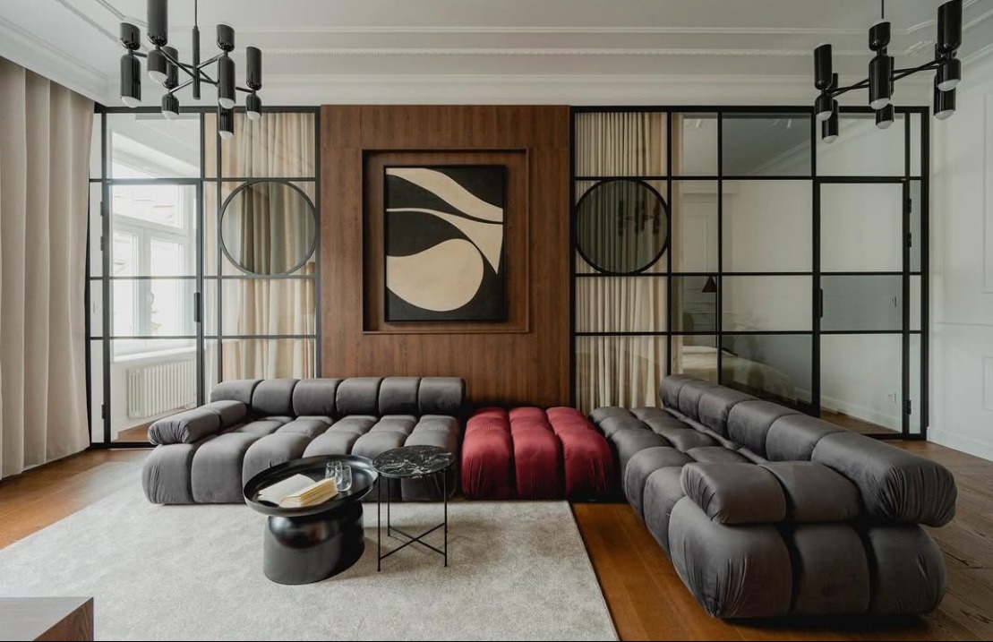 Nowoczesny salon z szarą sofą modułową, czerwonym pufem, stolikami kawowymi z marmurowymi blatami i czarną, industrialną ścianą działową z lustrami w okrągłych ramach.