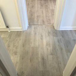 Kamil Rejmer tazz finishings - Widok z góry na jasną podłogę z paneli imitujących drewno, połączoną srebrną listwą progową między dwoma pomieszczeniami, widoczne białe ościeżnice drzwiowe.