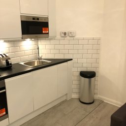 Kamil Rejmer tazz finishings - Nowoczesna kuchnia z białymi szafkami, czarnym blatem i białymi płytkami imitującymi cegłę, uzupełniona srebrnym koszem na śmieci i podłogą z paneli w odcieniach szarości.