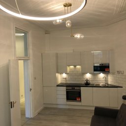 Kamil Rejmer tazz finishings - Nowoczesna kuchnia z białymi, błyszczącymi szafkami, czarnym blatem i białymi płytkami na ścianie, oświetlona okrągłą lampą sufitową i punktowymi światłami pod szafkami, z widocznym fragmentem...