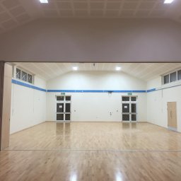 Kamil Rejmer tazz finishings - Sala gimnastyczna po odświeżeniu: jasne ściany z niebieskim akcentem, parkiet na podłodze, widoczne drzwi ewakuacyjne i okna pod sufitem, oświetlenie sufitowe.