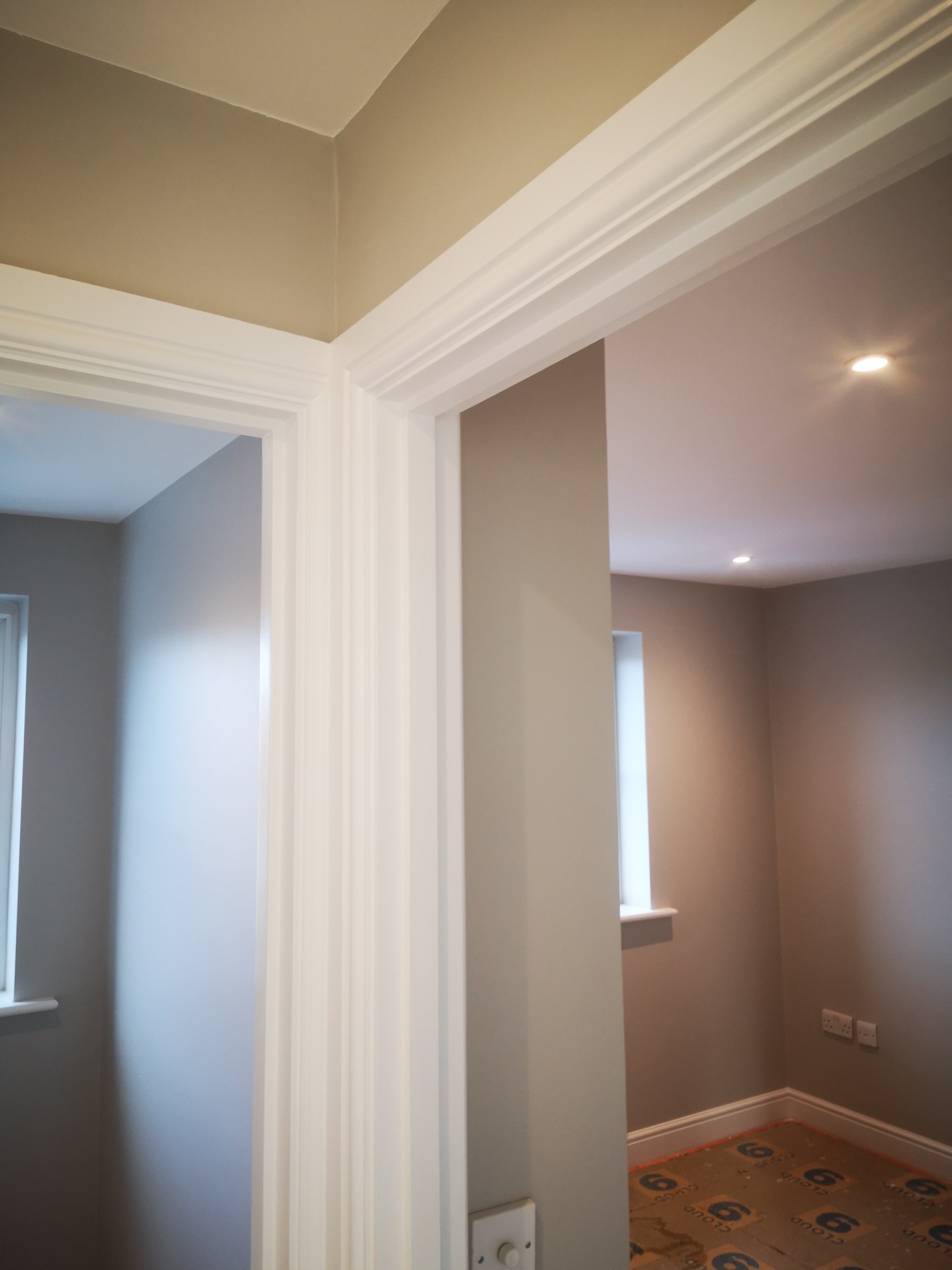 Świeżo pomalowane wnętrze z białymi, dekoracyjnymi listwami przypodłogowymi i ościeżnicami, widoczne gniazdka elektryczne i zabezpieczona podłoga.