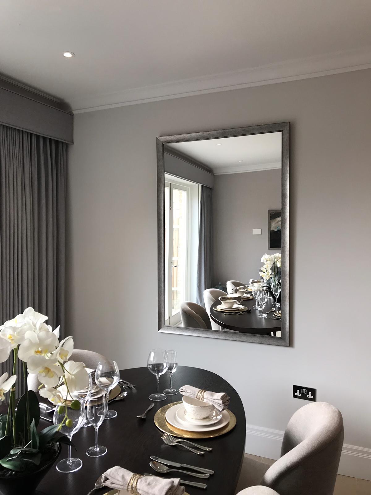 Elegancko nakryty stół w jadalni z ciemnego drewna, widoczny w lustrze na szarej ścianie. Zastawa stołowa z elementami złota, szklanki do wina, lniane serwetki. W tle szare zasłony i białe orchidee.