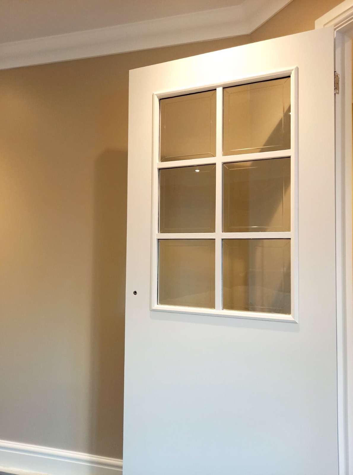 Białe drzwi wewnętrzne z sześcioma szybkami, częściowo otwarte, widoczne na tle beżowej ściany z białą listwą przypodłogową i sufitową.