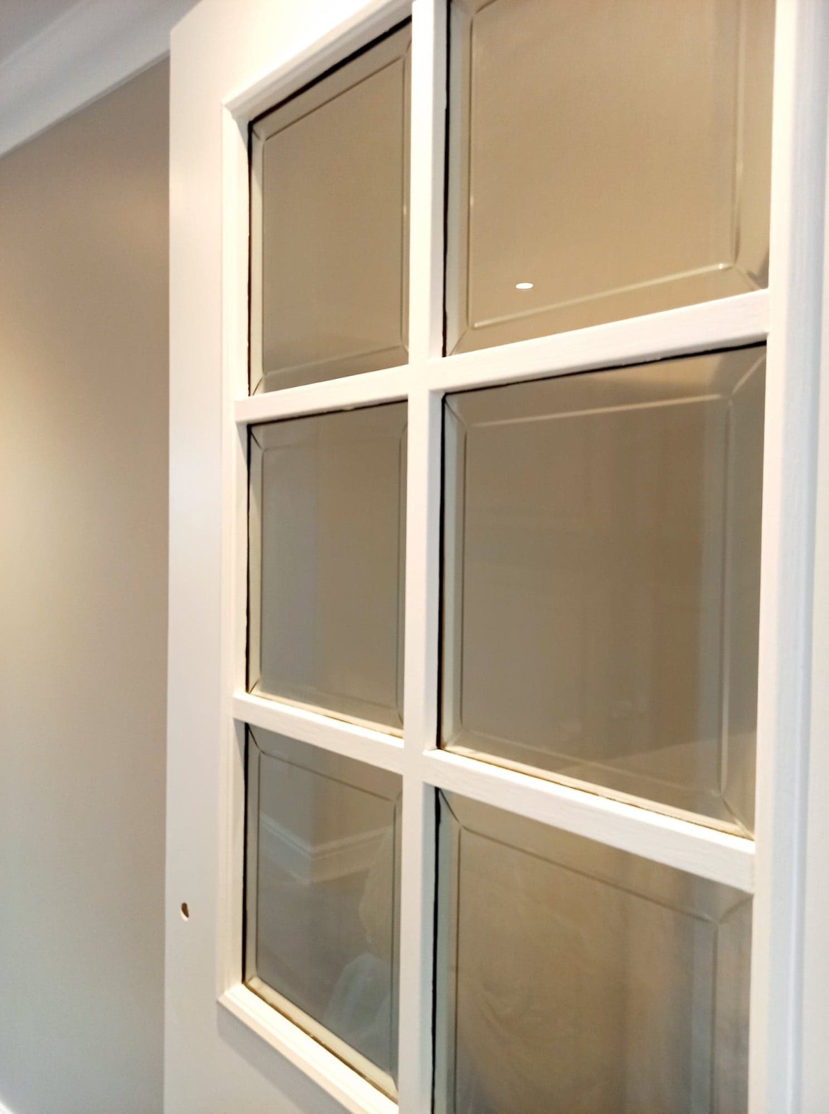 Białe, przeszklone drzwi wewnętrzne z sześcioma kwadratowymi szybami, widoczne z bliska, z delikatnym odbiciem wnętrza na powierzchni szkła.