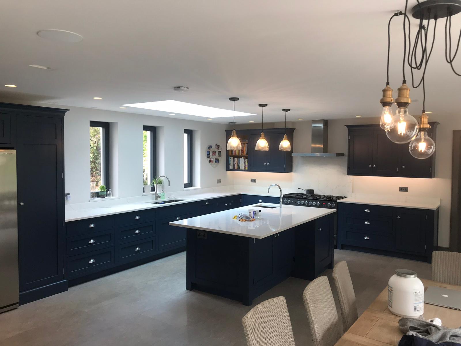 Nowoczesna, przestronna kuchnia w odcieniach granatu z białym blatem, wyspą kuchenną i oświetleniem punktowym oraz wiszącymi lampami z żarówkami Edisona.