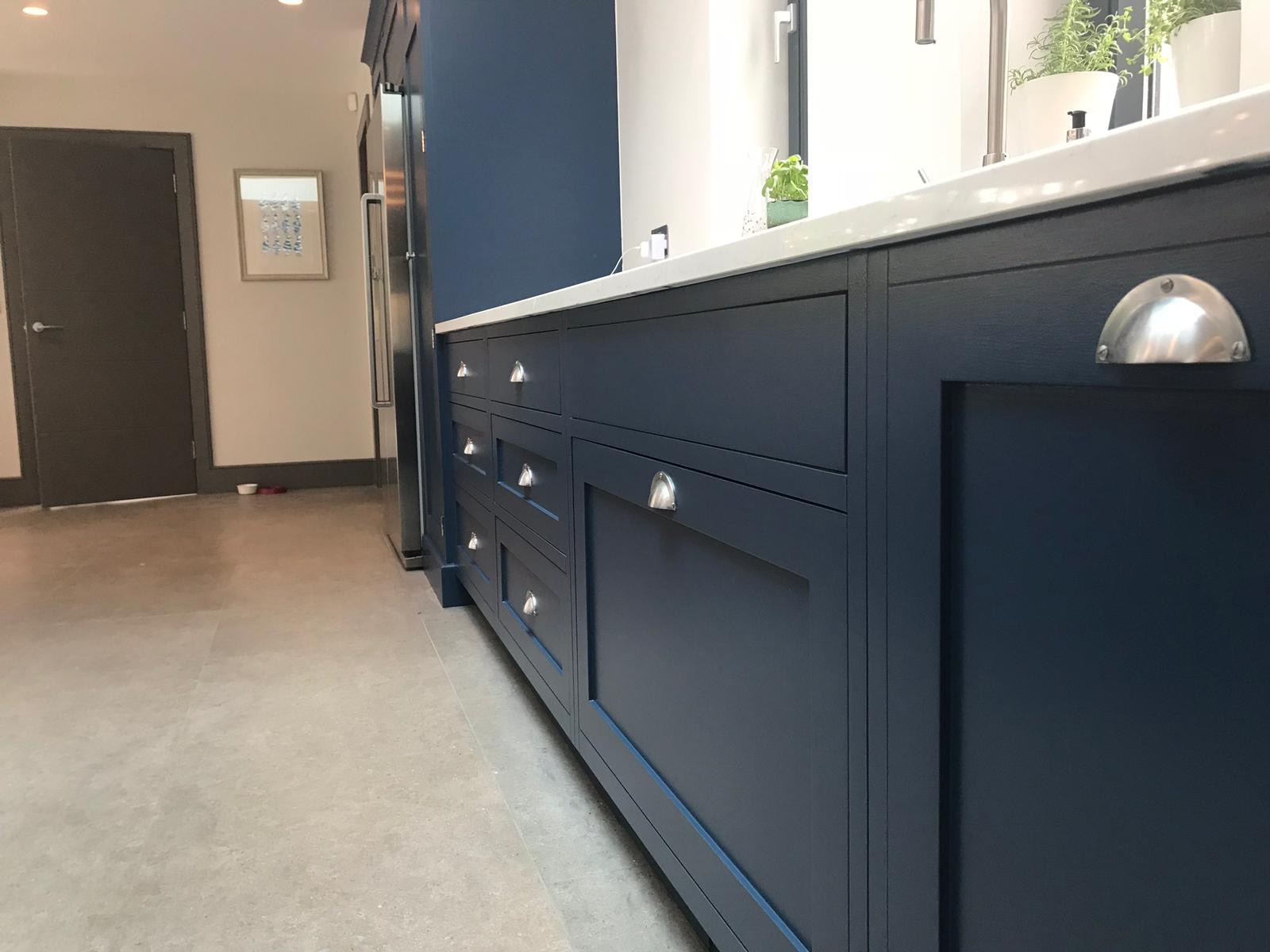 Granatowa zabudowa kuchenna z frezowanymi frontami, srebrnymi uchwytami typu muszelka i białym blatem, widok z niskiej perspektywy.