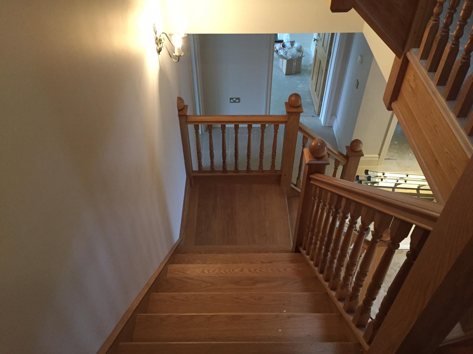Widok z góry na drewniane schody z balustradą, świeżo pomalowaną ścianą i widocznym w tle kartonem oraz rozłożoną drabiną, sugerującymi trwające prace.