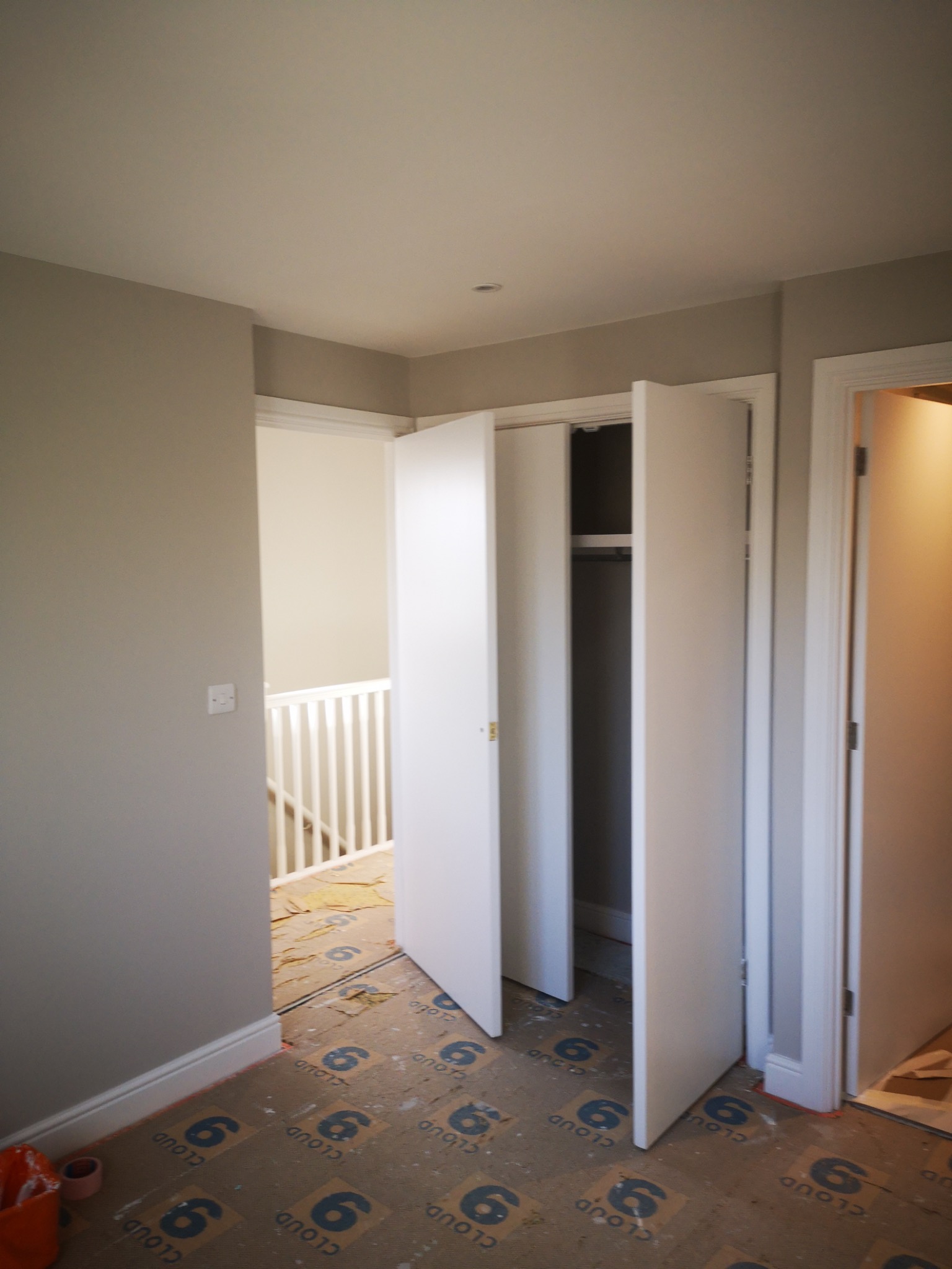 Wnętrze w trakcie prac wykończeniowych: widok na otwarte drzwi szafy wnękowej z białymi skrzydłami, podłoga zabezpieczona kartonem z nadrukiem, szare ściany, widoczny fragment klatki schodowej w tle.