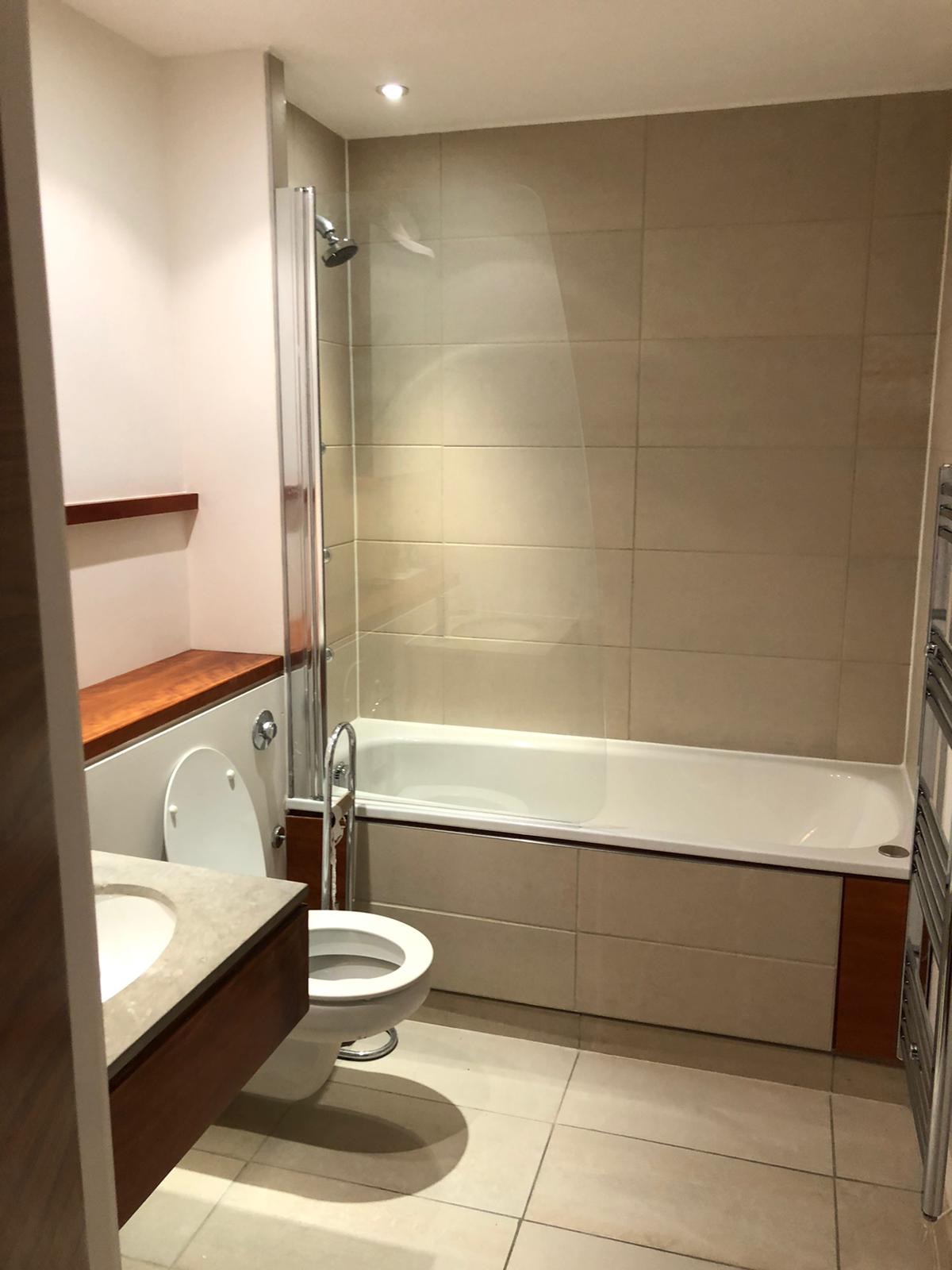 Nowoczesna łazienka z beżowymi płytkami, białą wanną z zabudową z elementami drewna, umywalką z kamiennym blatem i szklanym parawanem nawannowym.