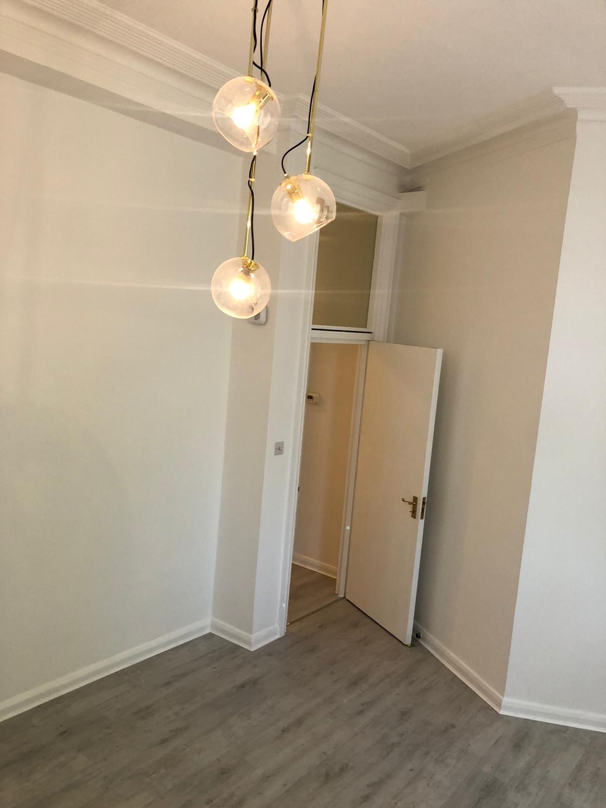 Jasne wnętrze po odświeżeniu, z trzema szklanymi lampami wiszącymi na złotych przewodach, widoczne listwy przypodłogowe i drzwi do kolejnego pomieszczenia.