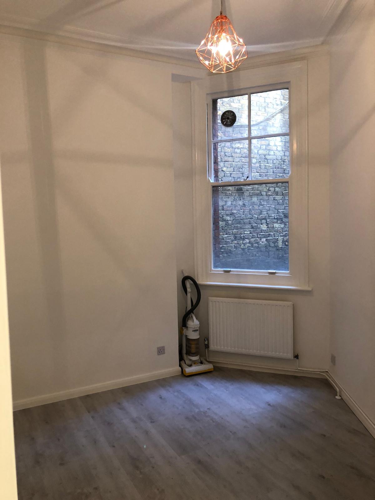 Pomalowane na biało wnętrze pokoju z szarą podłogą, widoczne okno z ceglanym murem za szybą, biały grzejnik i odkurzacz stojący przy ścianie.