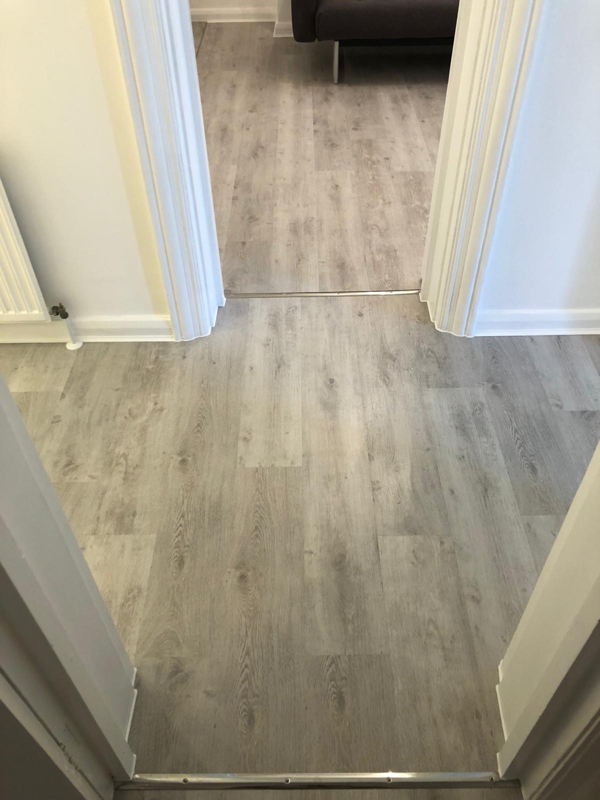 Widok z góry na jasną podłogę z paneli imitujących drewno, połączoną srebrną listwą progową między dwoma pomieszczeniami, widoczne białe ościeżnice drzwiowe.