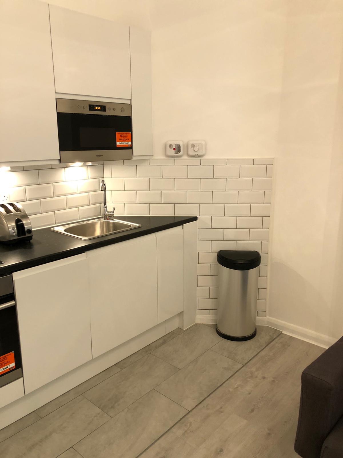 Nowoczesna kuchnia z białymi szafkami, czarnym blatem i białymi płytkami imitującymi cegłę, uzupełniona srebrnym koszem na śmieci i podłogą z paneli w odcieniach szarości.