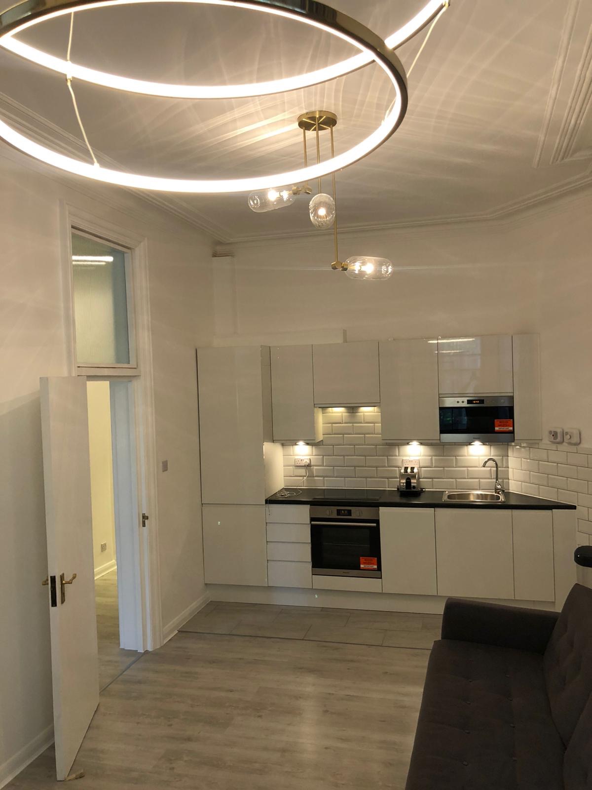 Nowoczesna kuchnia z białymi, błyszczącymi szafkami, czarnym blatem i białymi płytkami na ścianie, oświetlona okrągłą lampą sufitową i punktowymi światłami pod szafkami, z widocznym fragmentem...