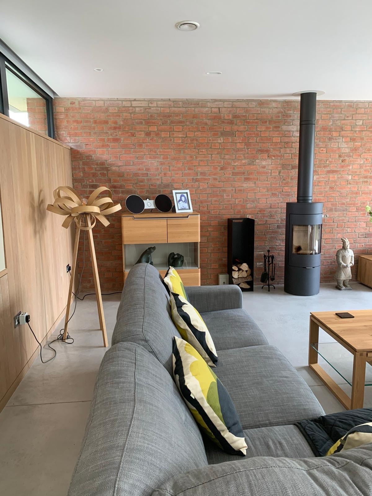 Jasne wnętrze salonu z ceglaną ścianą, szarą sofą z wzorzystymi poduszkami, czarnym piecem kominkowym i drewnianymi meblami, oświetlone naturalnym światłem z okna po lewej stronie.