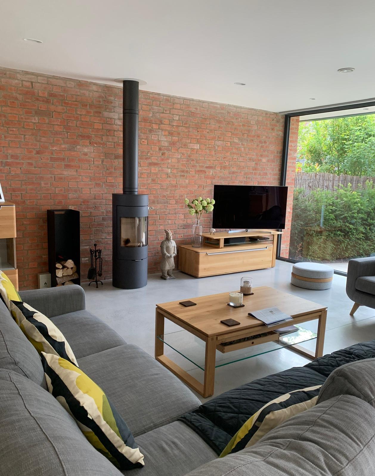 Salon z ceglaną ścianą, nowoczesnym piecem wolnostojącym, szarą sofą z wzorzystymi poduszkami i drewnianym stolikiem kawowym z szklaną półką.