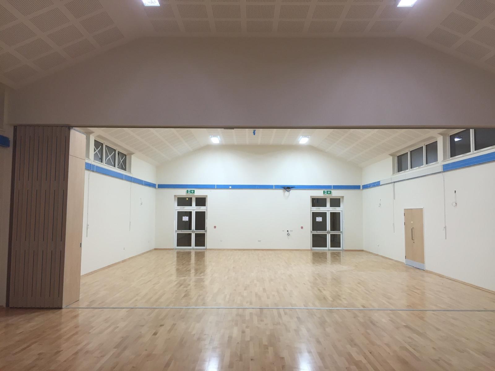 Sala gimnastyczna po odświeżeniu: jasne ściany z niebieskim akcentem, parkiet na podłodze, widoczne drzwi ewakuacyjne i okna pod sufitem, oświetlenie sufitowe.