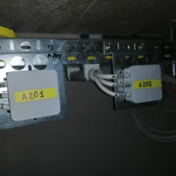 Dwa szare puszki instalacyjne z żółtymi etykietami 'A201' i 'A202' przymocowane do metalowej szyny na suficie, z widocznymi przewodami oznaczonymi numerami.