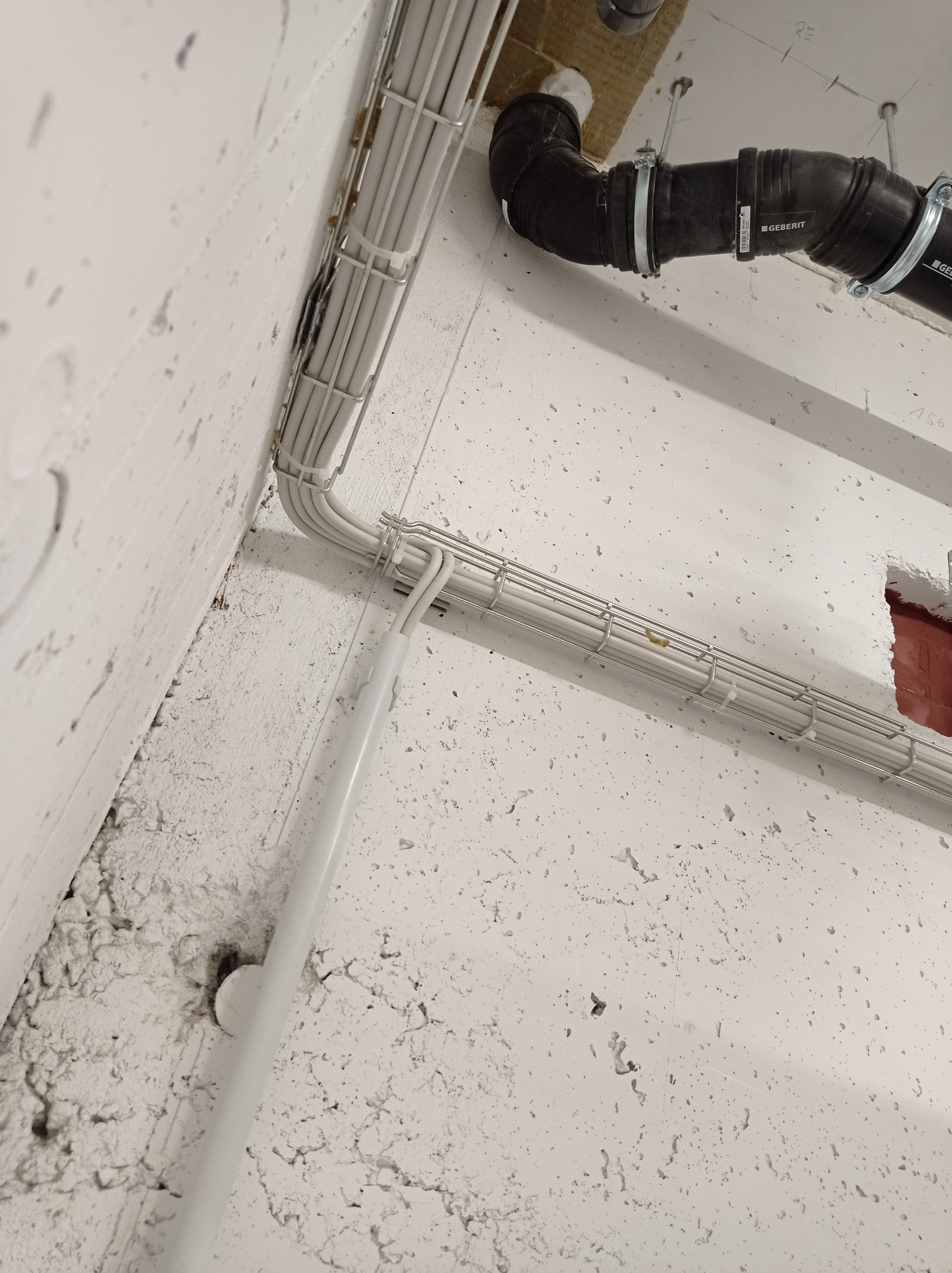 Białe kable elektryczne ułożone w metalowej korycie na surowej, białej ścianie, obok czarnej rury kanalizacyjnej z logo Geberit.