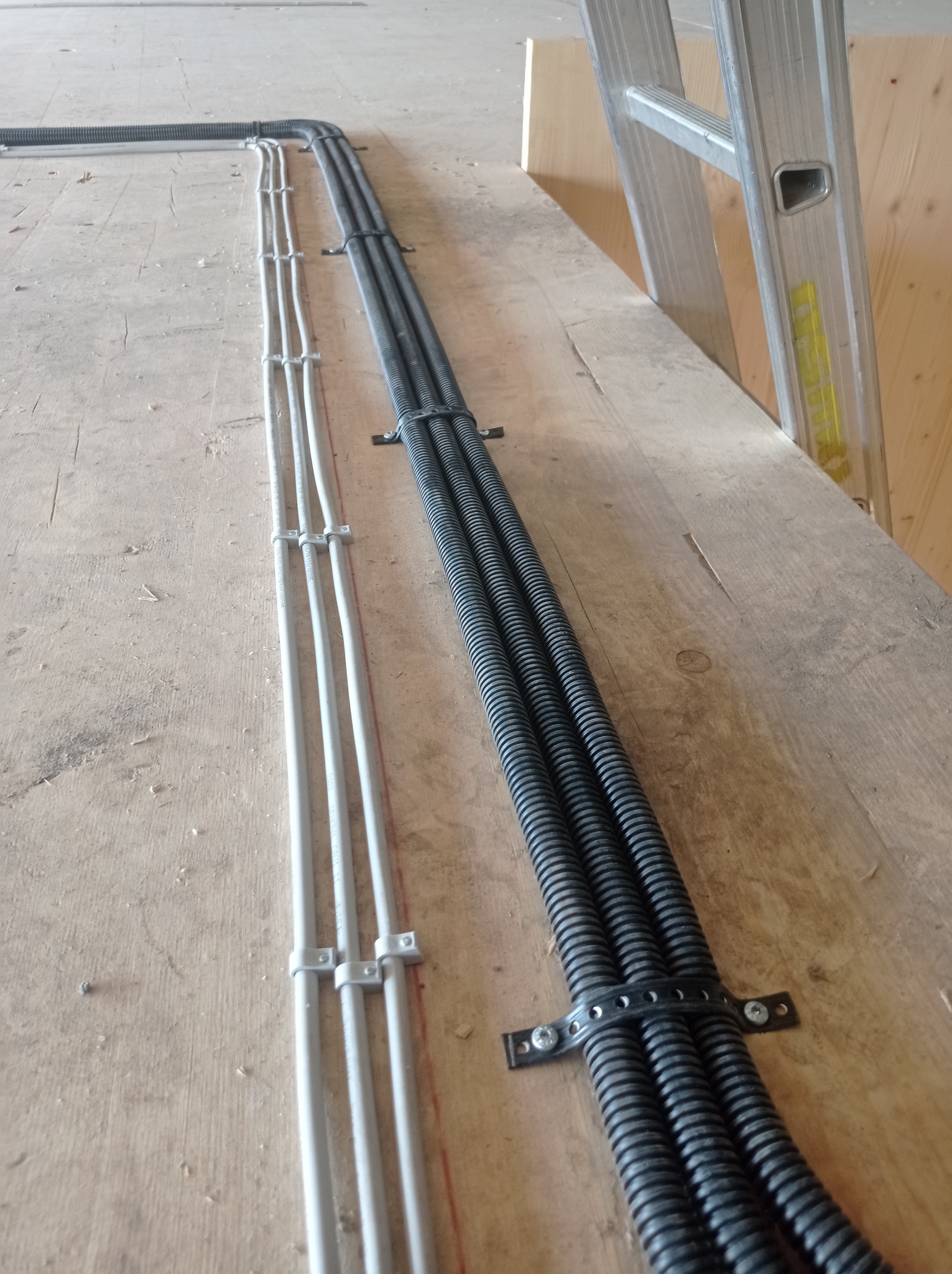 Ułożone równolegle kable elektryczne w peszlu i rurki PCV przymocowane do drewnianej powierzchni, widoczna drabina aluminiowa w tle.