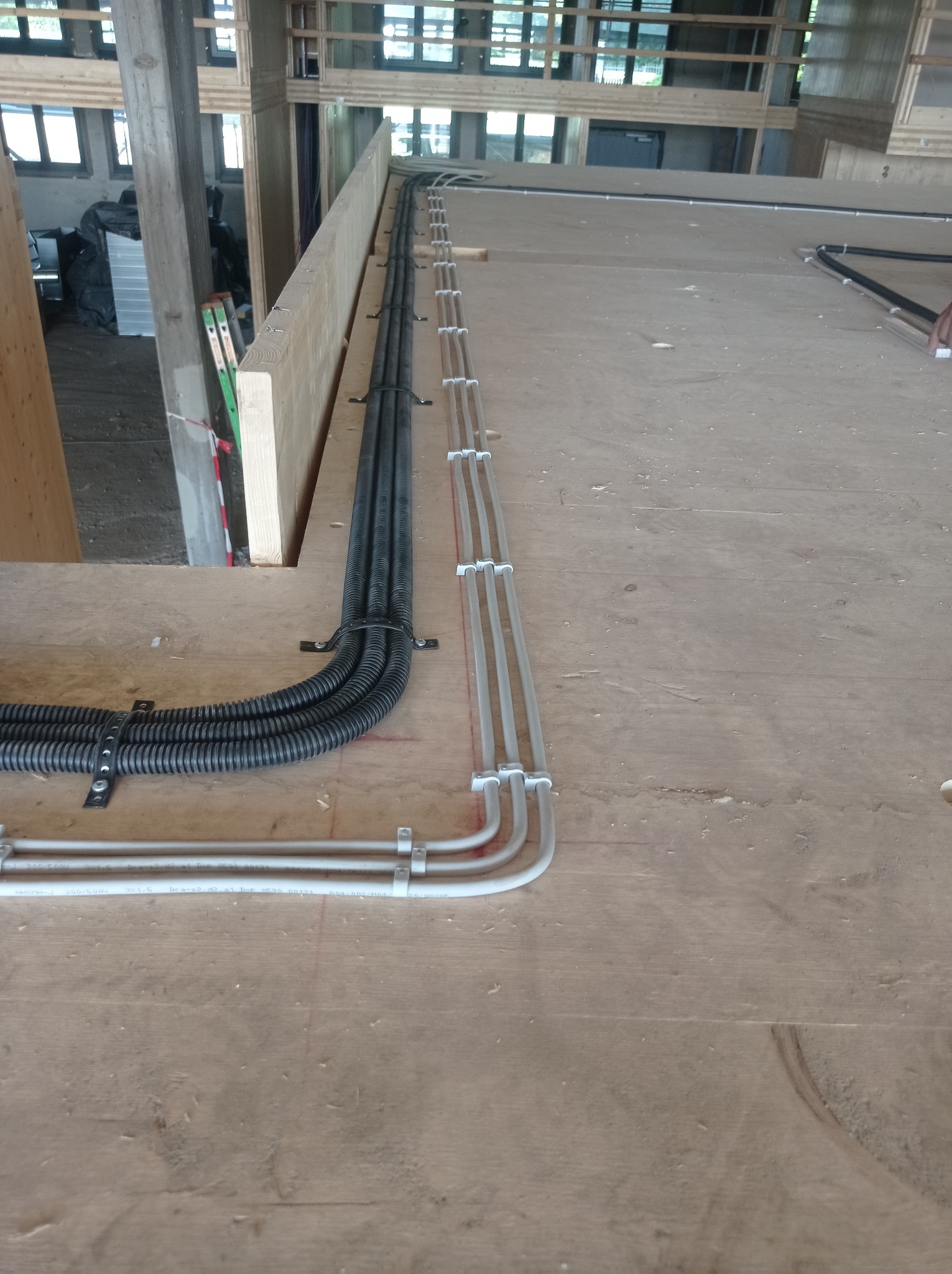 Rzędy przewodów elektrycznych, czarnych karbowanych i szarych gładkich, zamocowane do drewnianej konstrukcji budynku w trakcie budowy.