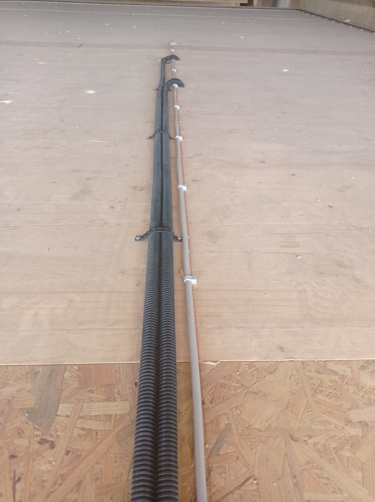 Dwa czarne karbowane kable elektryczne oraz rurka instalacyjna przymocowane do surowej płyty OSB za pomocą metalowych i plastikowych uchwytów.