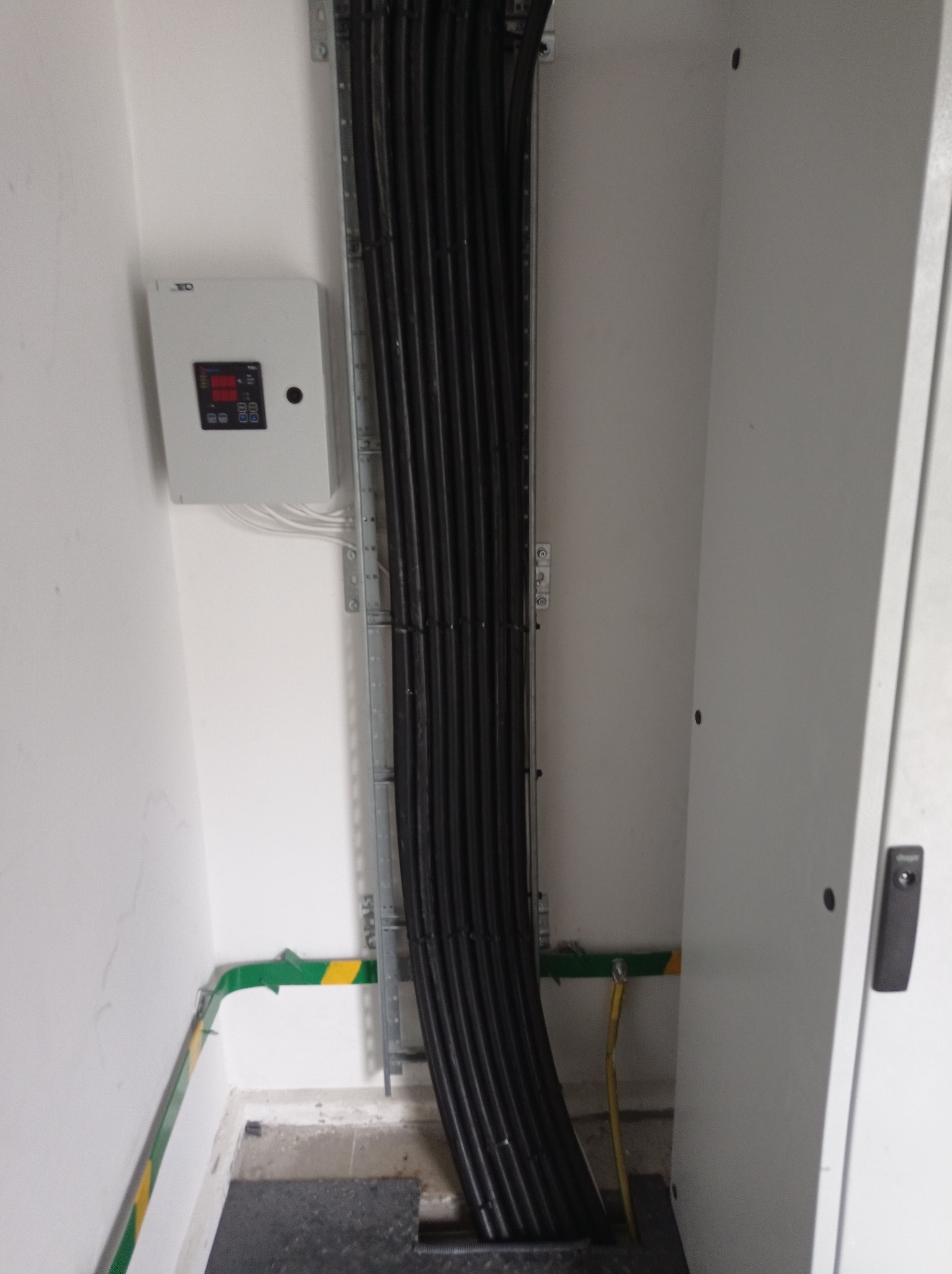 Pionowy układ kabli elektrycznych w korycie, z widocznym panelem sterowania i uziemieniem, wewnątrz białego pomieszczenia technicznego.