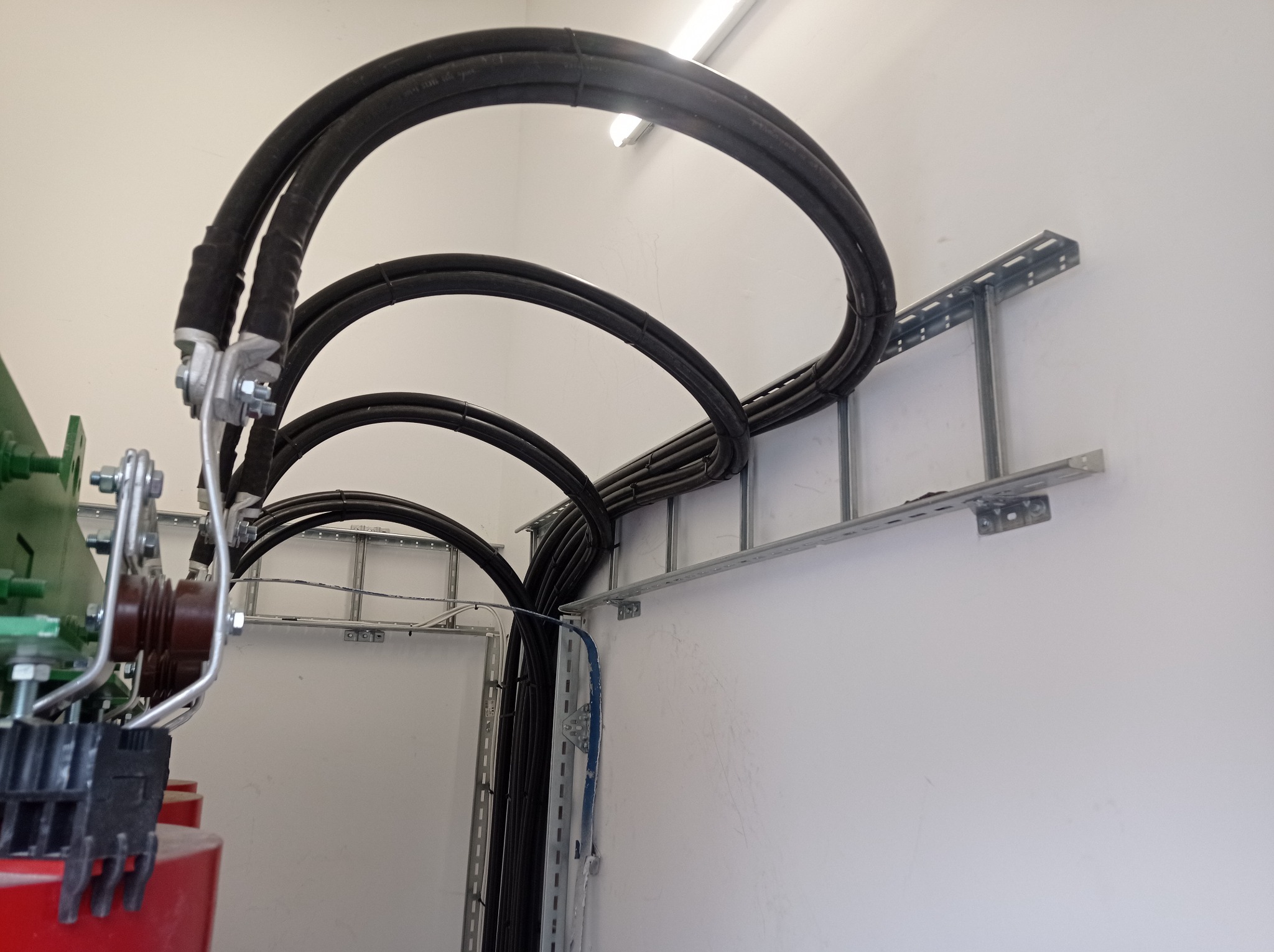 Grube kable elektryczne ułożone w pętle na metalowej drabince przy ścianie, widoczne złącza i mocowania.