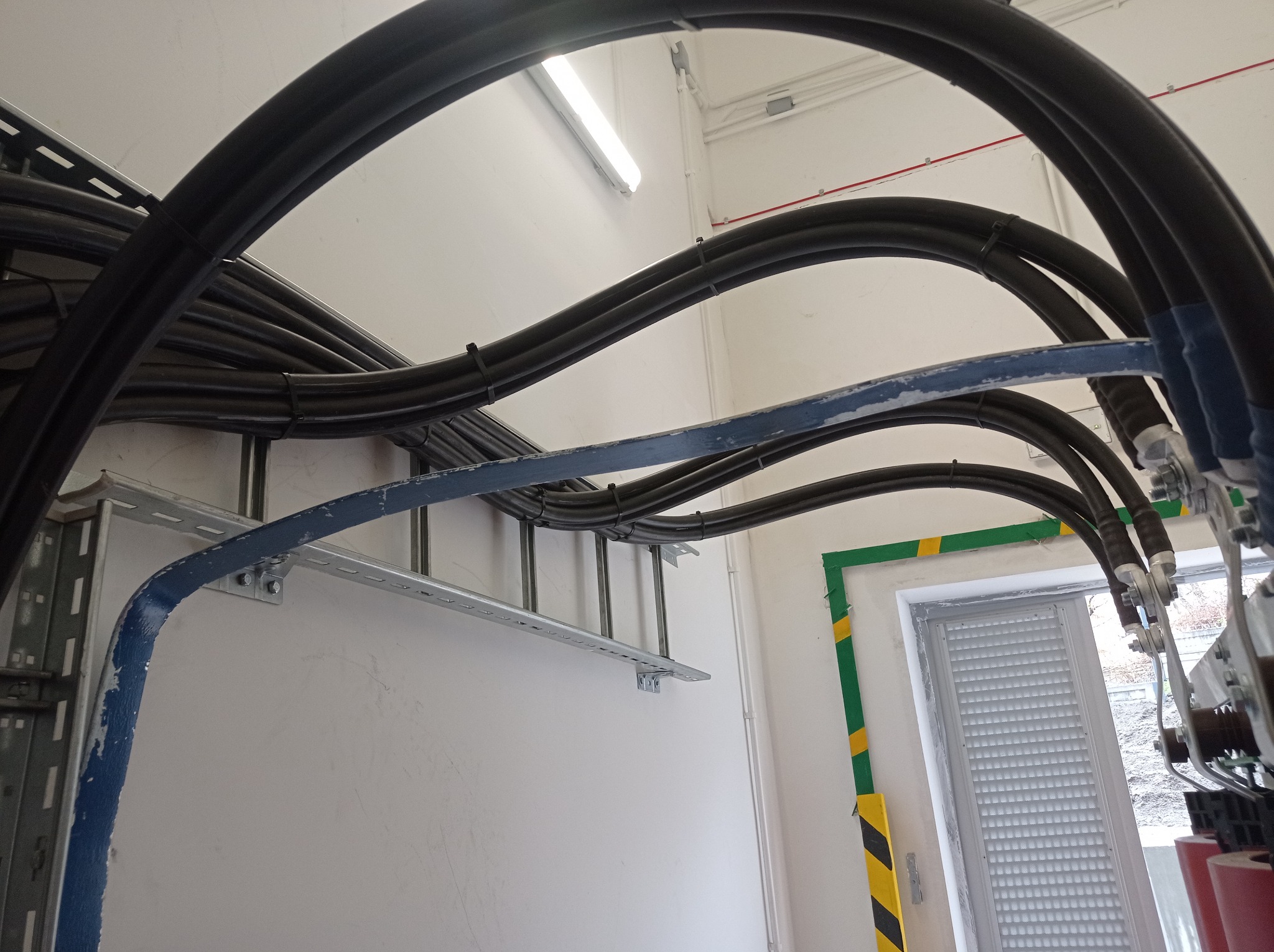 Układ kabli elektrycznych w czarnych peszlach, ułożonych na metalowej drabince przymocowanej do ściany, z widocznym oknem w tle.