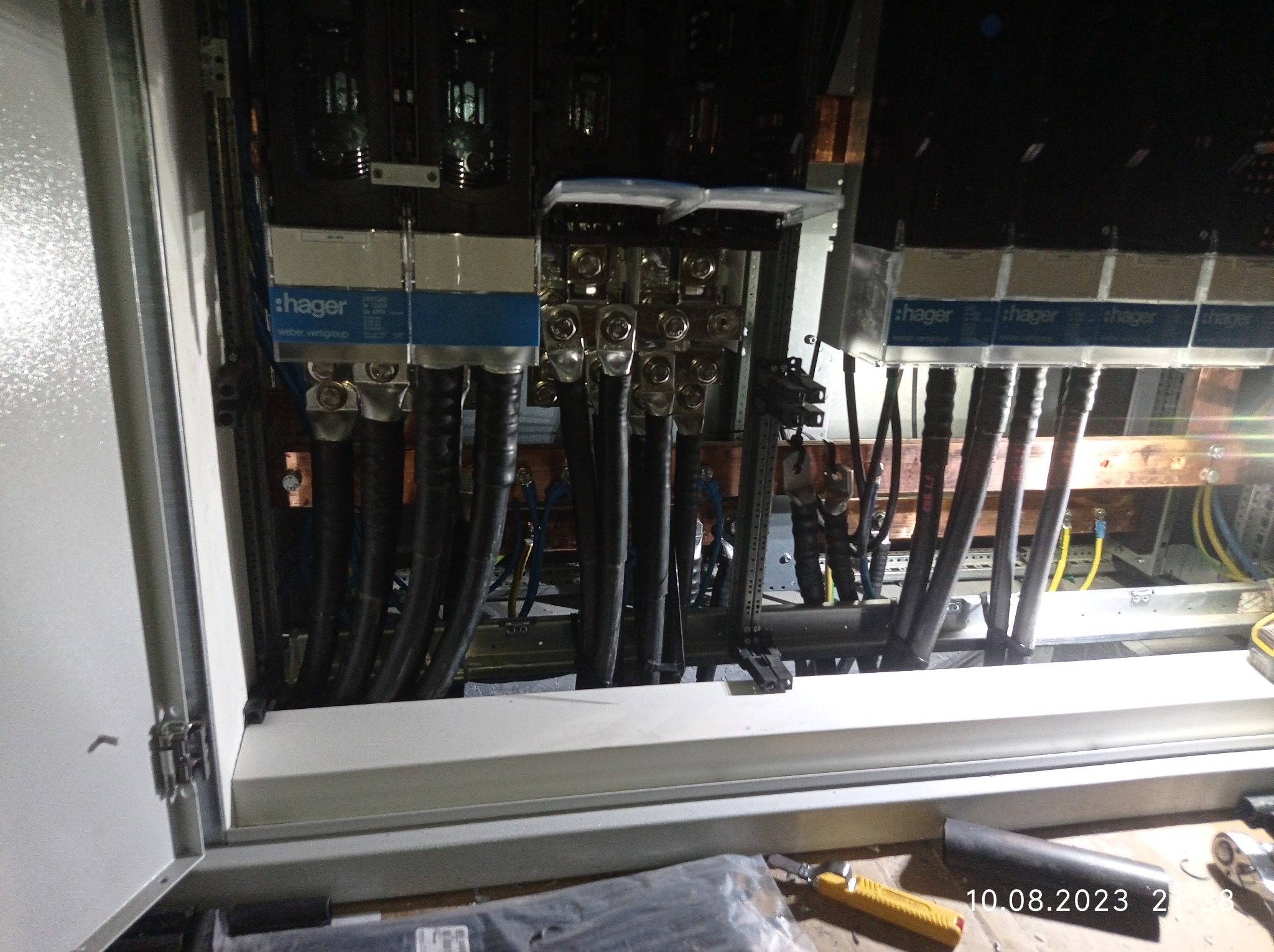 Otwarta szafa rozdzielcza z widocznymi przewodami, złączkami i elementami marki Hager, oświetlona od wewnątrz, z narzędziami leżącymi na dole.