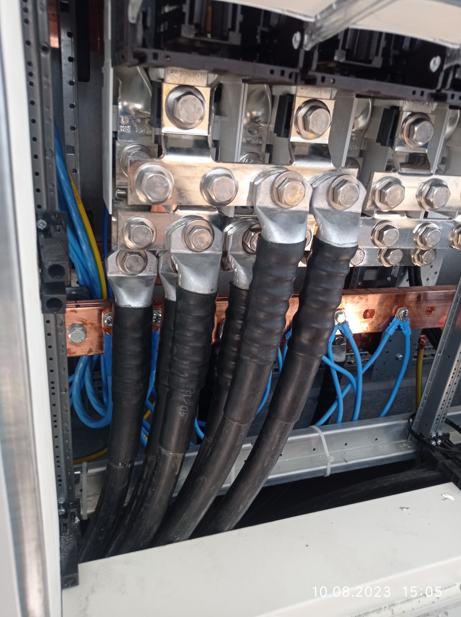 Szczegółowe ujęcie wnętrza rozdzielnicy elektrycznej z grubymi kablami zasilającymi zakończonymi srebrnymi końcówkami, podłączonymi do miedzianych szyn zbiorczych za pomocą śrub; widoczne także...