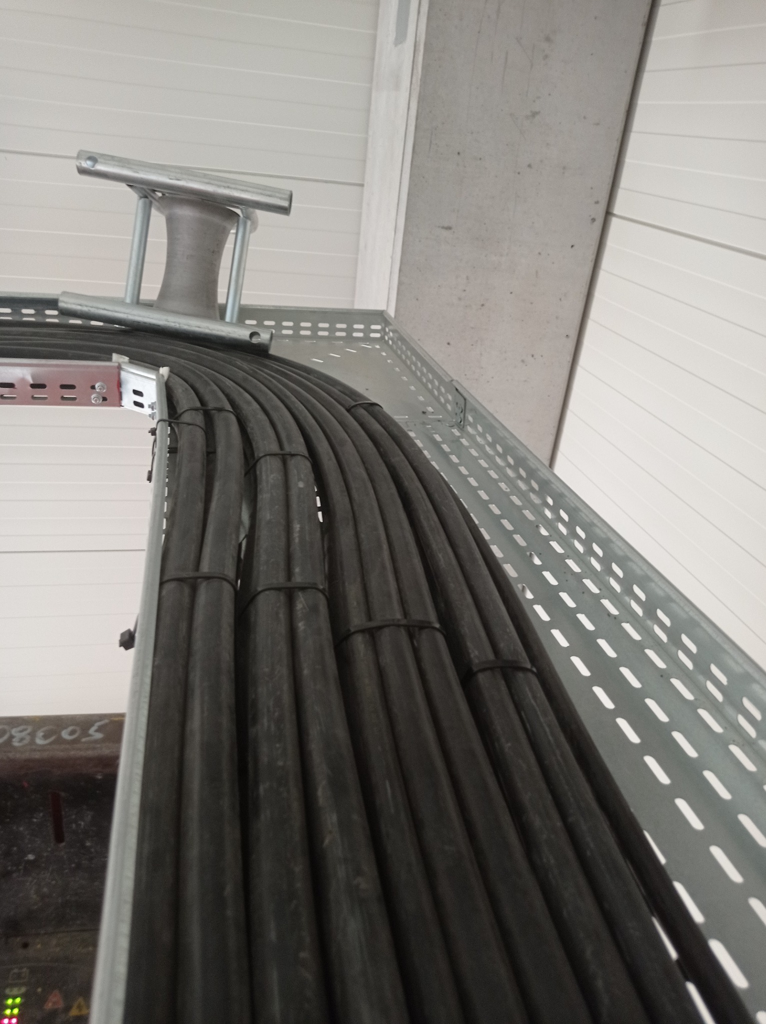 Rząd czarnych kabli elektrycznych ułożonych w perforowanej, metalowej korycie kablowej, zabezpieczonych czarnymi opaskami, z widocznym uchwytem montażowym i fragmentem betonowej ściany w tle.