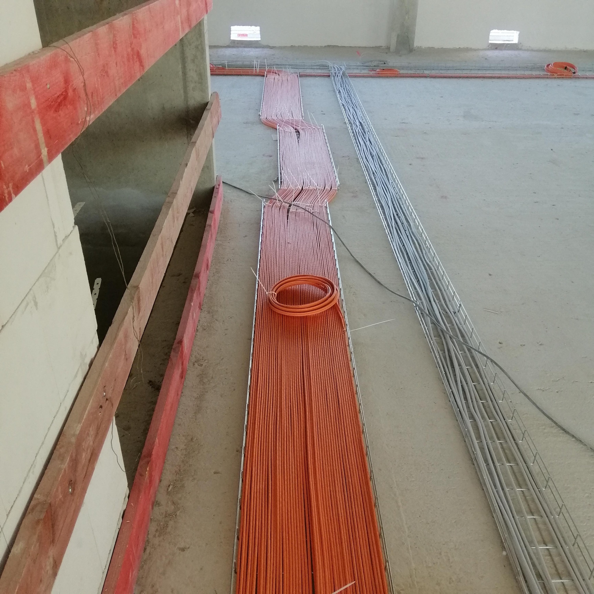Pomarańczowe przewody elektryczne ułożone równolegle w metalowej korycie kablowej na surowym betonowym podłożu w trakcie układania instalacji elektrycznej w nowym budynku.