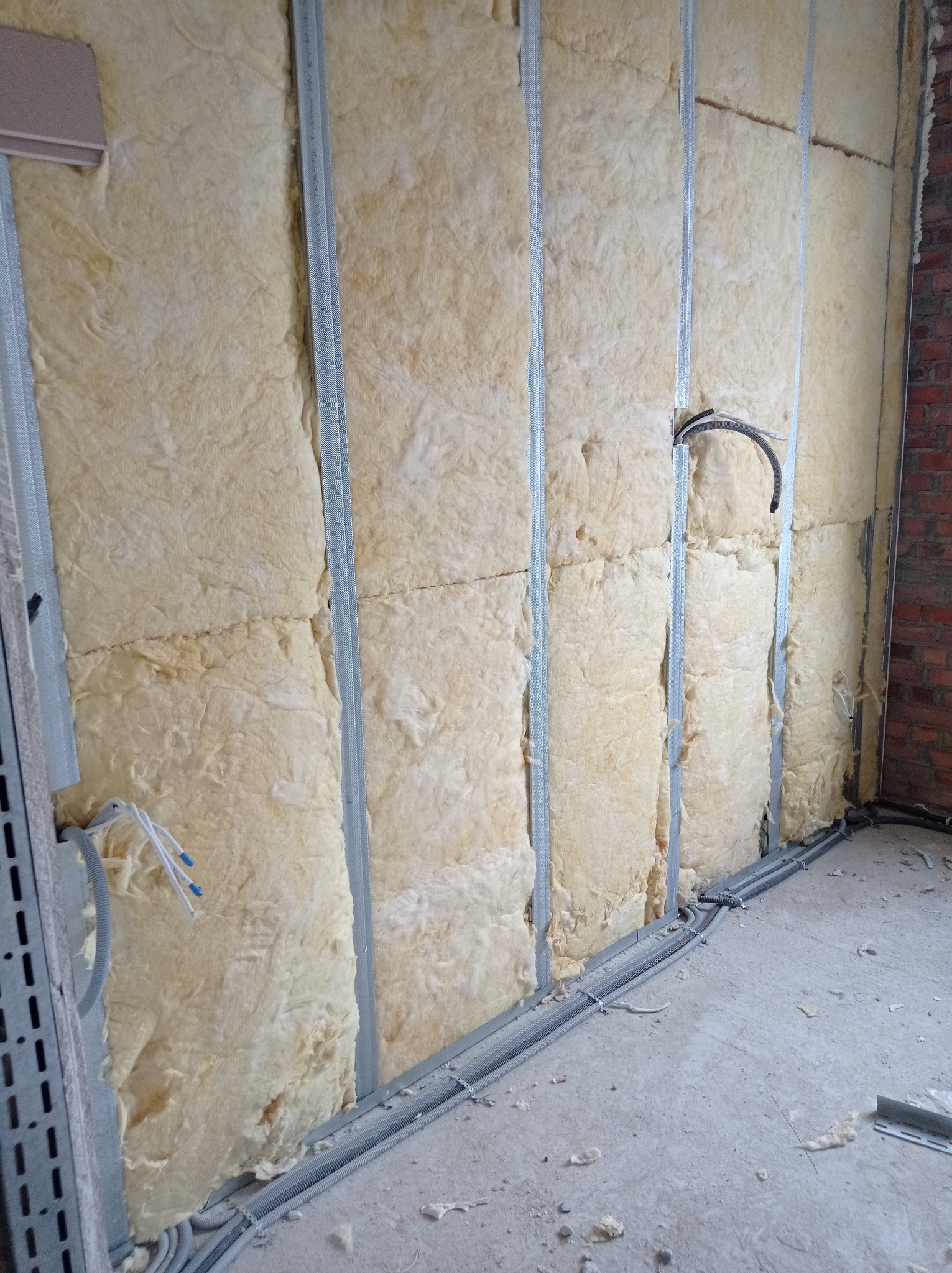 Ściana w trakcie izolacji wełną mineralną, widoczne metalowe profile i instalacja elektryczna w peszlach.