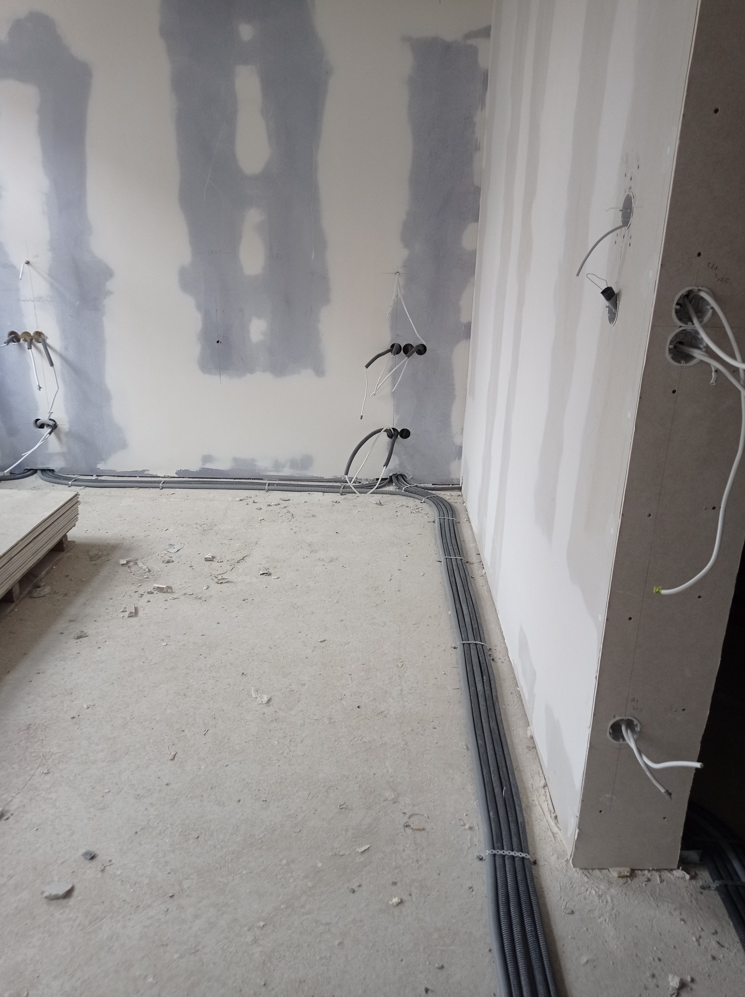 Wnętrze w trakcie prac wykończeniowych: ściany z płyt kartonowo-gipsowych z widocznymi przewodami elektrycznymi w peszlach i puszkach instalacyjnych, ułożonymi wzdłuż podłogi, oraz stos płyt...