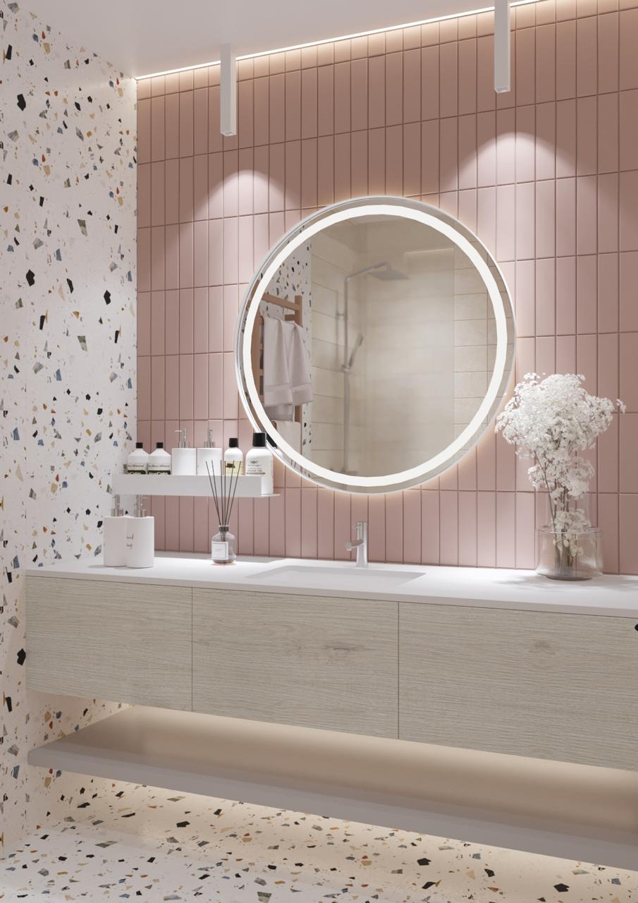 Nowoczesna łazienka z okrągłym lustrem podświetlanym LED, jasnymi płytkami w odcieniach różu i lastryko, drewniana szafka pod umywalką i szklany wazon z białymi kwiatami.