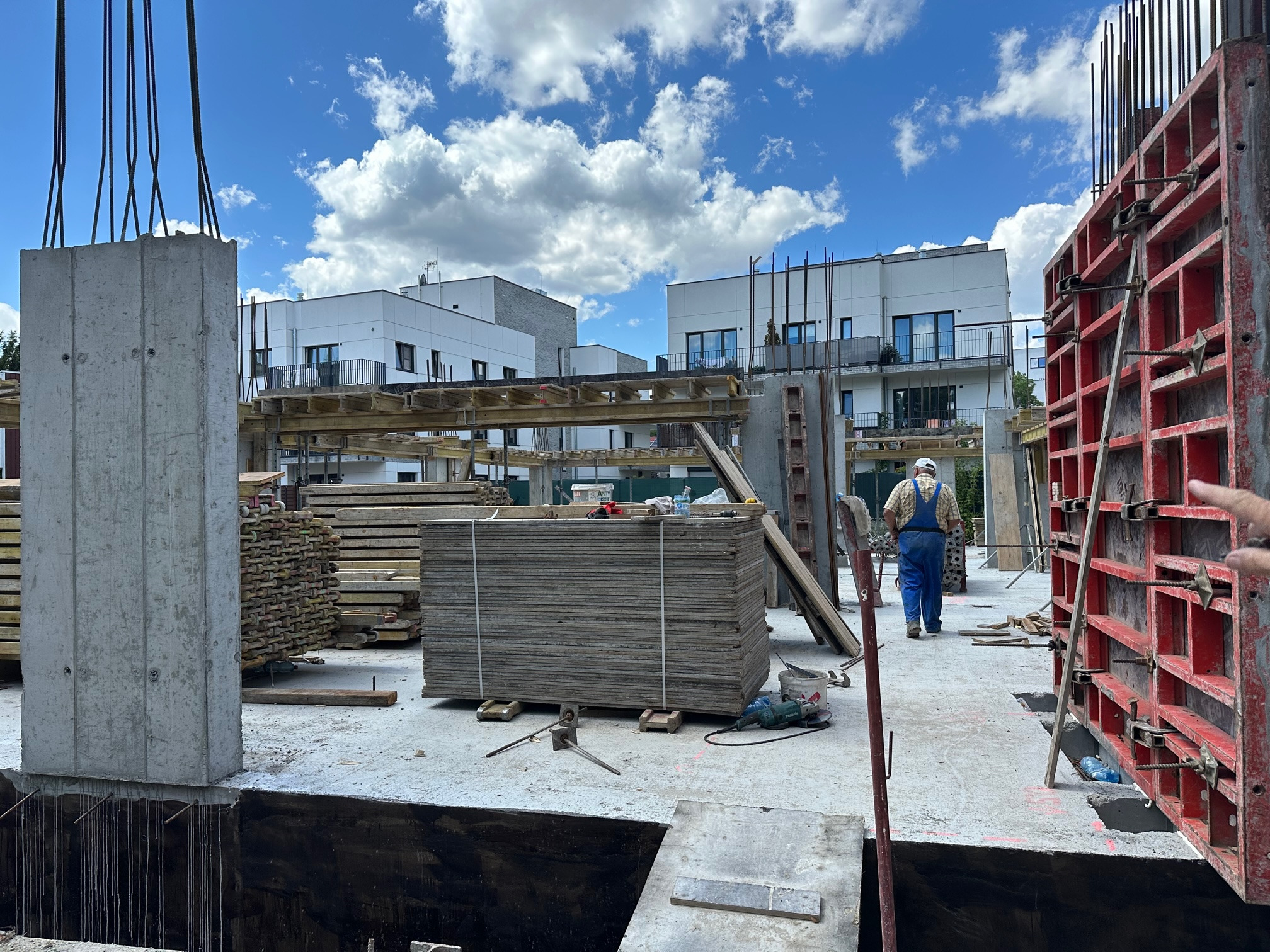 Wznoszenie budynku: widok na plac budowy z betonowym słupem z wystającymi prętami zbrojeniowymi, deskowaniem ścian, stertą drewnianych płyt oraz pracownikiem w oddali, na tle nowoczesnych budynków...