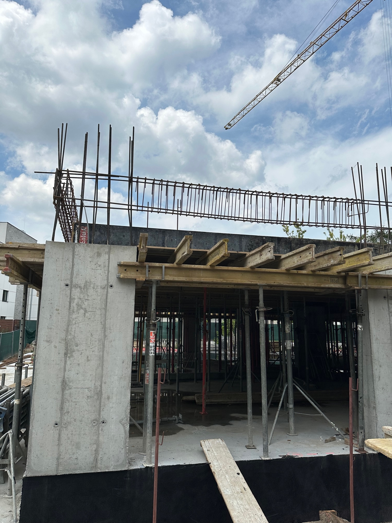 Wznoszenie żelbetowej konstrukcji w Warszawie: szalunki, zbrojenie i podpory widoczne na placu budowy, w tle błękitne niebo z chmurami i fragment żurawia.