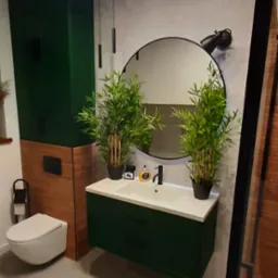 Nowoczesna łazienka z zielonymi akcentami: wisząca toaleta, szafka pod umywalkę w kolorze butelkowej zieleni, okrągłe lustro z oświetleniem punktowym, rośliny w doniczkach i szara podłoga.