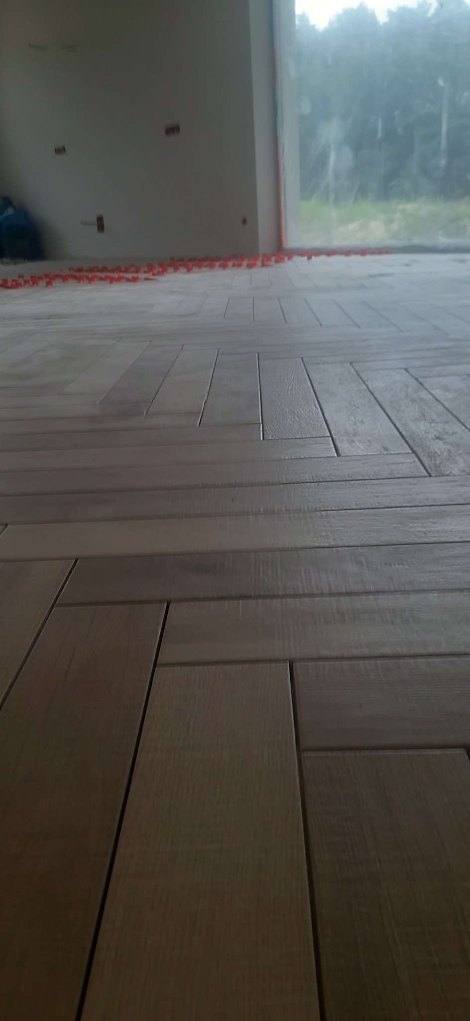 Układanie paneli podłogowych w jodełkę, widoczne czerwone kliny dystansowe przy ścianie, surowy stan wykończenia ścian.