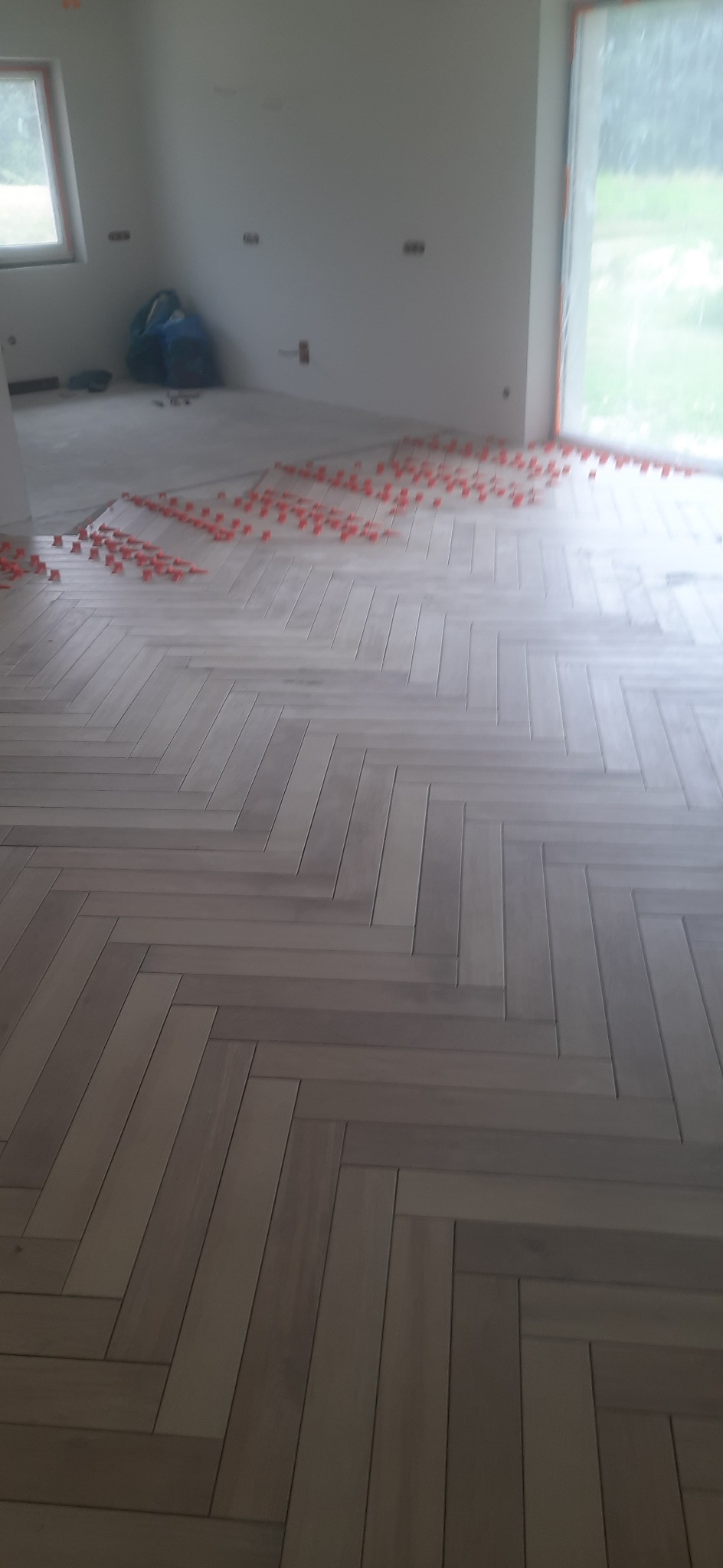 Układanie jasnoszarych paneli podłogowych w jodełkę w nowym pomieszczeniu, widoczne plastikowe kliny dystansowe i worki budowlane w tle.