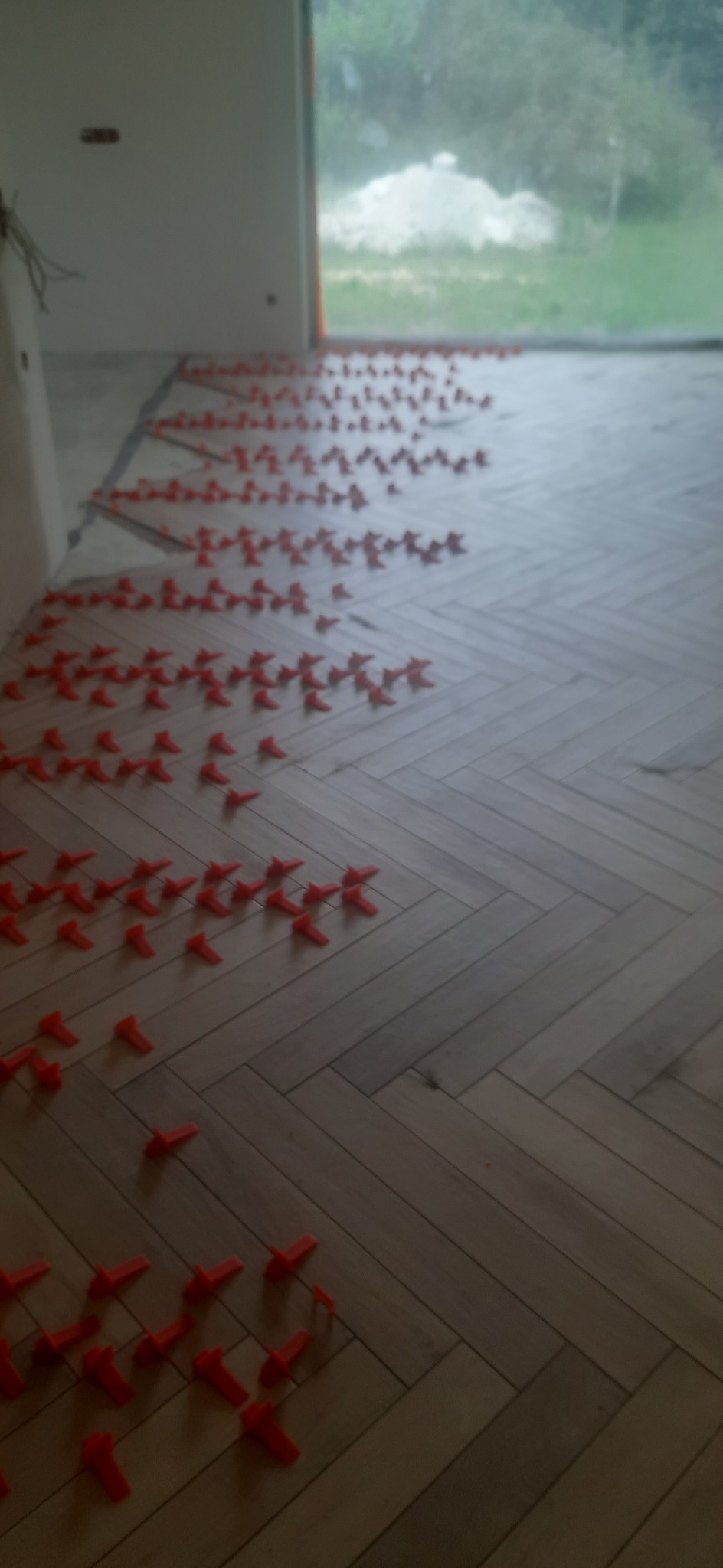 Ułożona podłoga z paneli w jodełkę, z widocznymi czerwonymi klipsami poziomującymi, okno w tle.