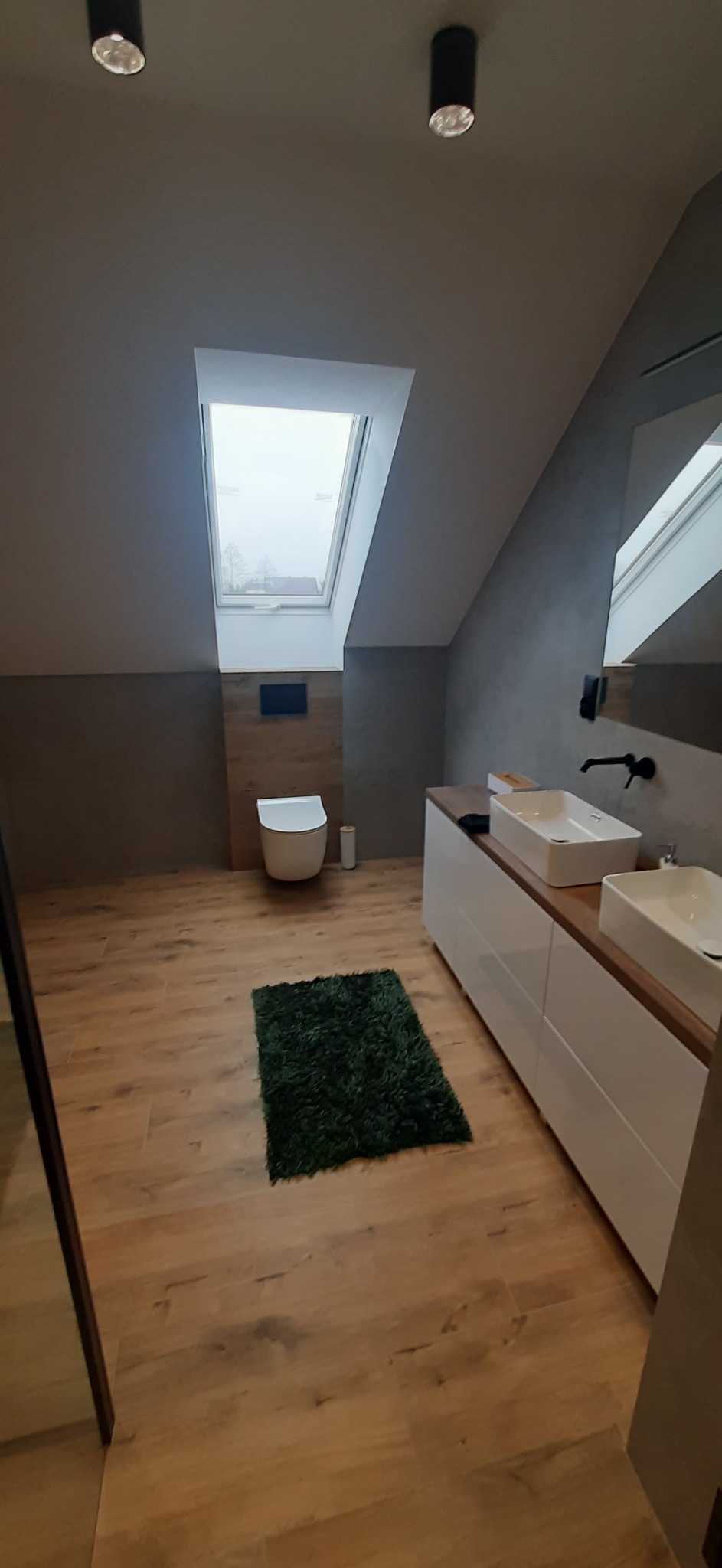 Nowoczesna łazienka na poddaszu z drewnianą podłogą, oknem dachowym i dwoma umywalkami nablatowymi na białej szafce, uzupełniona o ciemnozielony dywanik.