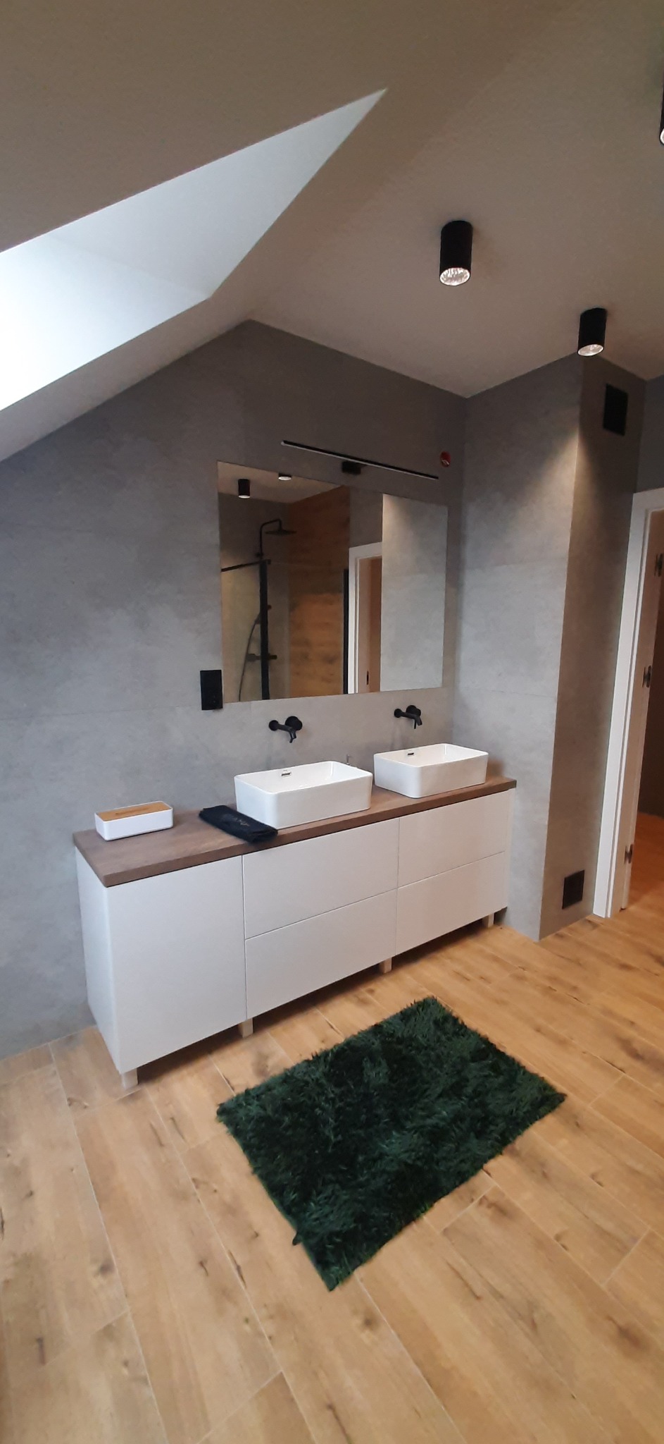 Nowoczesna łazienka na poddaszu z dwoma umywalkami nablatowymi, szarą ścianą z efektem betonu, drewnianym blatem i zielonym dywanikiem.