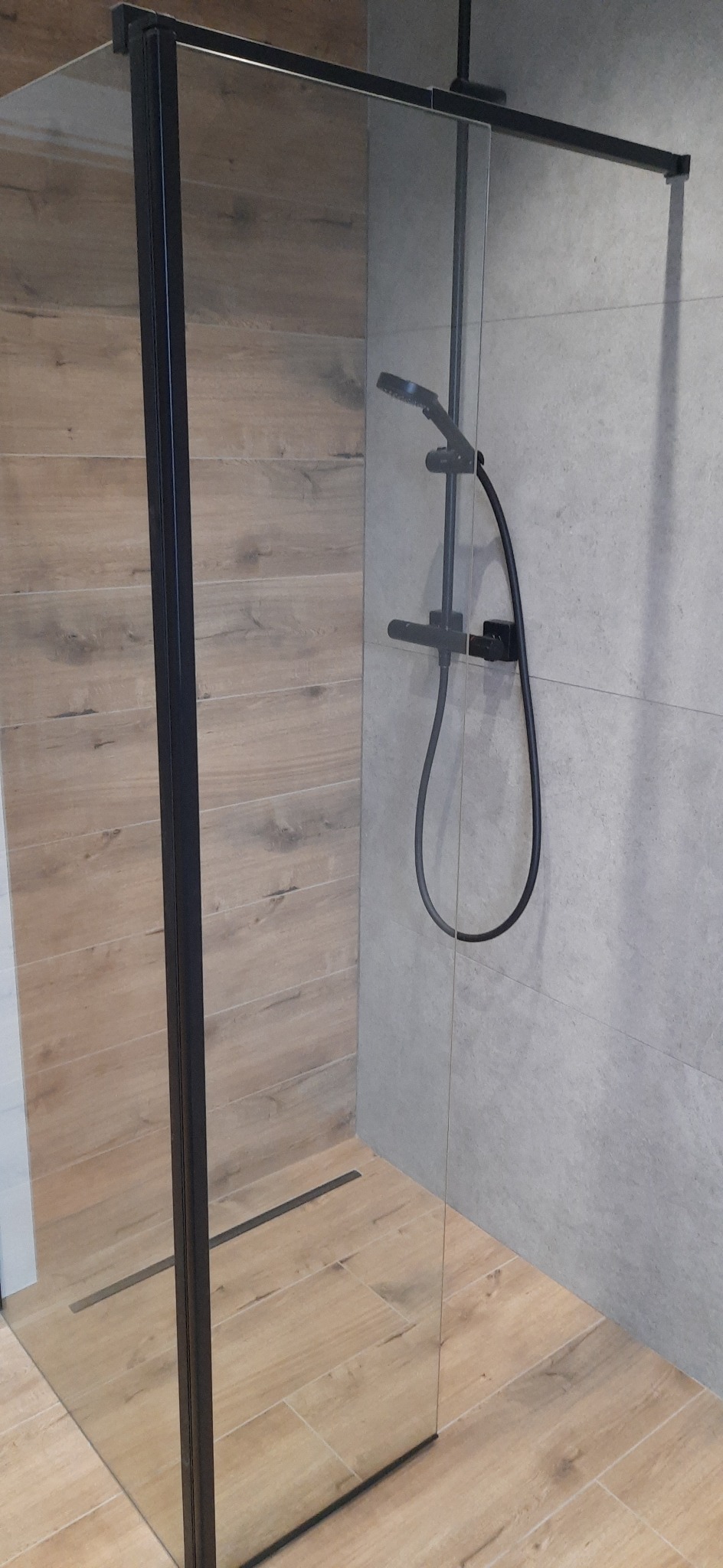 Nowoczesna kabina prysznicowa walk-in z czarną armaturą i odpływem liniowym, wykończona płytkami imitującymi drewno i szarym kamieniem.