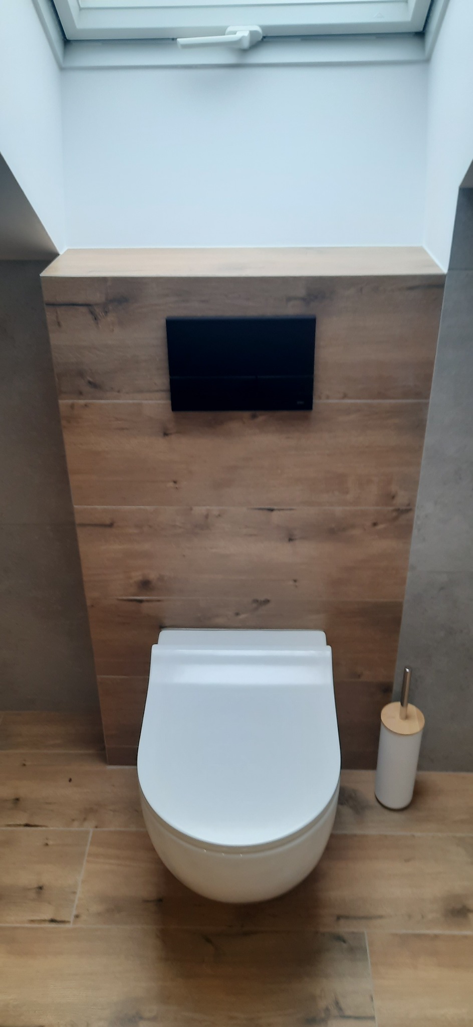 Nowoczesna toaleta podwieszana z czarnym przyciskiem spłukującym na tle drewnopodobnej ściany, oświetlona światłem z okna dachowego.