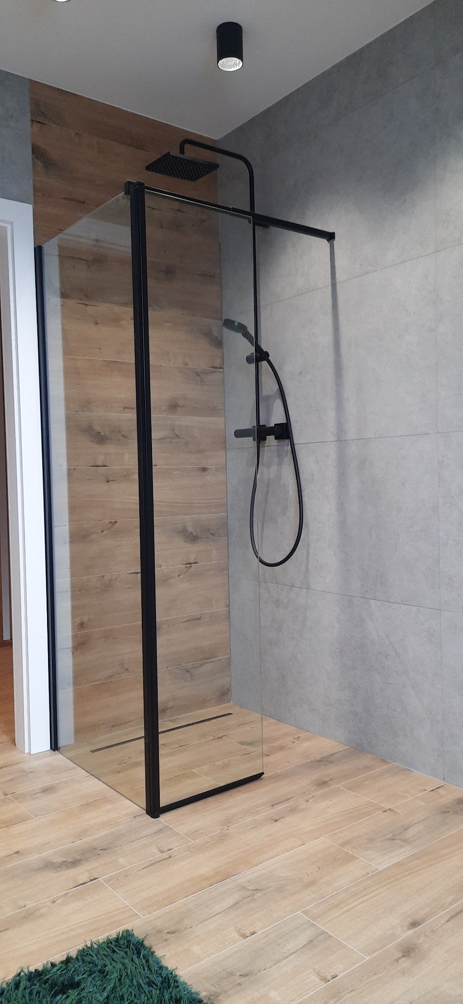 Nowoczesna kabina prysznicowa typu walk-in z czarnymi profilami, deszczownicą i baterią, na podłodze imitującej drewno, z zielonym dywanikiem.