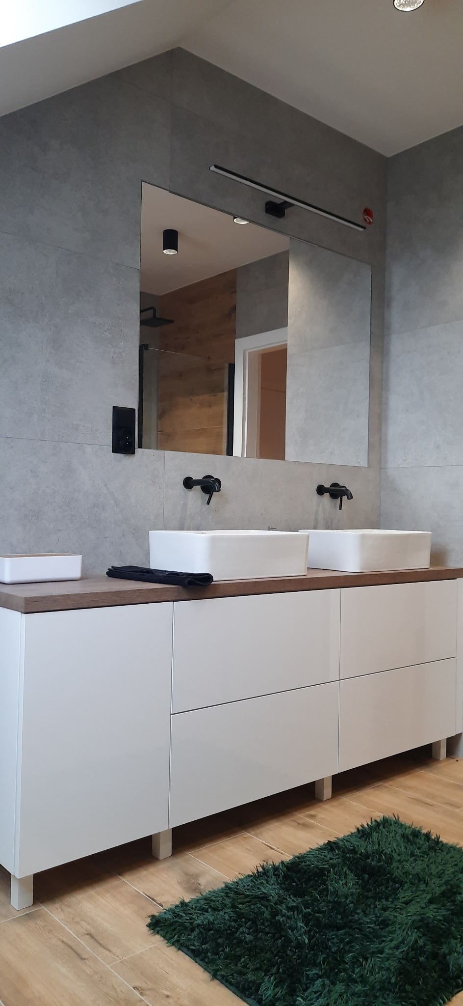 Nowoczesna łazienka z podwójną umywalką nablatową, czarną armaturą i szarymi płytkami na ścianach, uzupełniona o zielony dywanik.