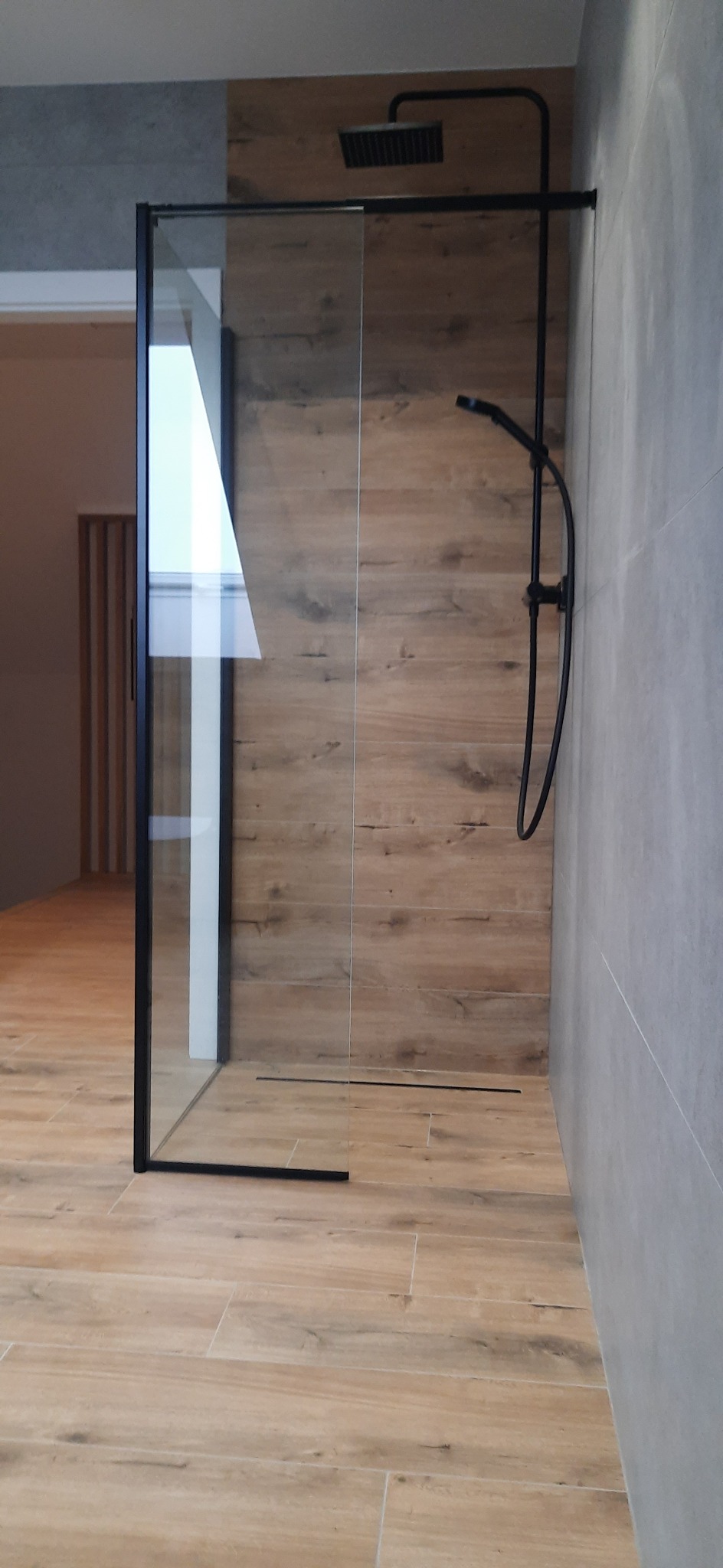 Nowoczesna kabina prysznicowa walk-in z czarną armaturą, drewnopodobne płytki na ścianie i podłodze, szklana ściana z czarną ramą, widoczny odpływ liniowy.